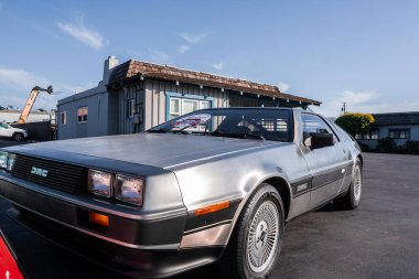 Paslanmaz çelikten DeLorean DMC 12 martı kanatlı kapılar Monterey 'de park halinde, öğleden sonra ışığı uzun gölgeler oluşturur, kama biçimi ve türbin tekerlekleri öne çıkar.
