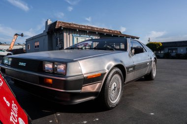 Paslanmaz çelikten bir DeLorean DMC 12 Monterey, California 'da asfalt bir arsada bulunuyor. Öğleden sonra ışığı uzun gölgeler bırakır. Martı kanadı dikişleri, tekerlekler ve DMC rozetini gösterir..