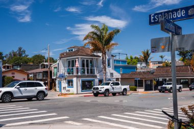 Palmiye ağaçları, pastel cepheler ve arabalar Capitola, California 'da Esplanade ve Stockton' da bir yaya geçidinde, gün ortası ışığı altında, taretli beyaz ve deniz mavisi bir binayla..