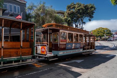 Powell ve Hyde pikabındaki bir San Francisco tramvayı gün ortası ışığı, kestane rengi ve krema, cilalanmış ahşap, Ghirardelli Meydanı için reklamlar, ağaçlar ve yakınlardaki evlerde oturur..