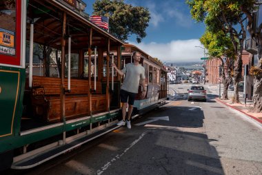 San Francisco teleferiğinin dış basamağında, körfez, alçak sis, çelik raylar, tuğla binalar ve Amerikan bayrağı görünür bir şekilde limana doğru inen bir adam var..