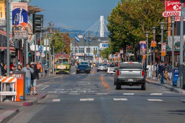 Arabalar San Francisco 'daki Embarcadero' ya giderken yayalar deniz ürünleri dükkanlarının önünden geçiyor. Körfez Köprüsü kuleleri açık gün ışığında pankart ve rıhtımların ötesine yükseliyor.