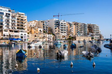 Sliema, Malta limanı sakin bir limanın üzerinde modern apartmanlar ve kireçtaşı binalar gösteriyor. Küçük tekneler şamandıralarla sallanır, kırmızı vinç işaretleri gelişir, ışık kıtır kıtır.