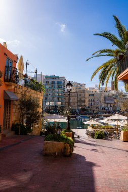 Sliema, Malta 'daki güneşli limanda kafeler, beyaz şemsiyeler, palmiye, botlar ve modern blokların yakınındaki kireçtaşı cepheleri, berrak öğleden sonra ışığı, deniz mavisi suyu, sosyal gezinti alanları gösteriliyor..