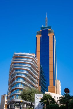 Portomaso Business Tower 'ın gündüz manzarası ve St. Julian' s, Malta 'daki kıvrımlı daireler. Mavi cam çekirdek, kule, kafes balkonları, palmiye ağaçları, marina işaretleri, kalın renkler..