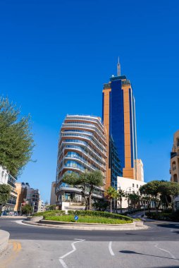 Parlak bir gün, budanmış ağaçlar ve peyzajlı bir ada, uzun bir turuncu ve mavi kule kulesi ve Tigne Yarımadası yakınlarında eğimli bir cam bina olan Sliema kavşağını gösteriyor..