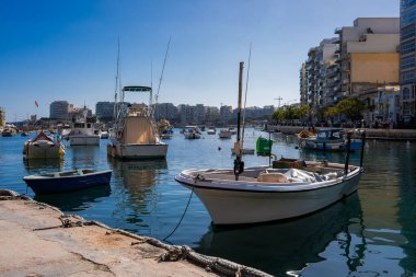 Sliema, Malta 'daki bir taş rıhtımının orada geleneksel balıkçı tekneleri ve el sanatları bataklığı. Durgun su direkleri yansıtırken, yükselen otellerin ve dairelerin ortası gezinti alanı..
