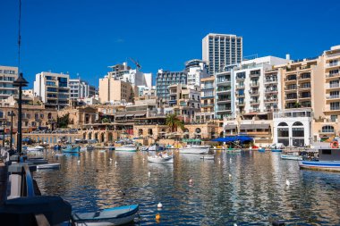 Sunlit Sliema, Malta, gün ortasında Balluta Körfezi yakınlarında şamandıralar, yansıtıcı su, kemerli taş depoları, palmiye ağaçları ve modern oteller tarafından sallanan renkli balıkçı tekneleri gösteriyor..