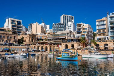 Dalgalanan yansımalar, kireçtaşı atari salonları, modern kuleler, palmiye ağaçları ve gündüz ışığı altında kafe teraslarıyla, Malta, Sliema 'da renkli luzzu tekneleri sallanıyor..