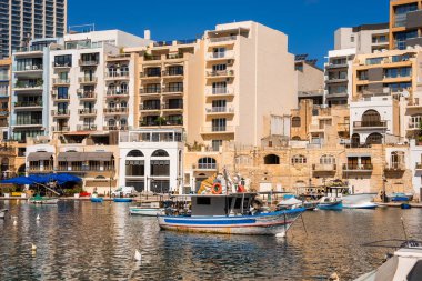 Malta 'nın Sliema limanında renkli tekneler sallanıyor. Kremalı taş daireler ve kemerli ön cepheler gibi. Gün ortası ışığı ve berrak su tekneleri ve balkonları yansıtıyor.