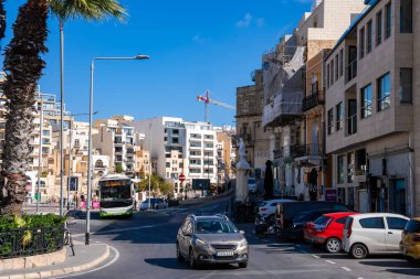 Sliema 'da gündüz görüşü, Malta' da kıvrımlı bir kıyı yolu, Malta otobüsü ve arabaları, Malta balkonlu kireçtaşı binalar, yüksek bir palmiye ve yukarıda kırmızı bir vinç göstermektedir..