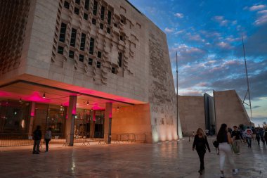 Valletta, Malta, Parlamento Binası 'nda delinmiş kireçtaşı ekranlar ve pembe ışık altı bir giriş var. İnsanlar asfalt meydanı geçerek mavi ve gül alacakaranlığında şehir kapısına doğru gidiyorlar..