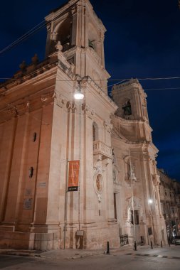 Valletta, Malta 'daki sıcak kireçtaşı kilise cephesi alacakaranlıkta parlıyor. İkiz çanlar, balkonlar, oyuklar ve boş caddenin üzerinde yumuşak lambaların altında duran oval pencere..