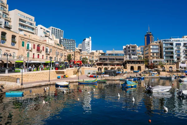 Sliema, Malta 'da renkli luzzu tekneleri, kemerli taş oyuklar, kafeler ve yüksek bir kule yakınında dinleniyor. Gün ortası ışığı ve sakin su, pastel cepheleri ve modern düzlükleri yansıtır..