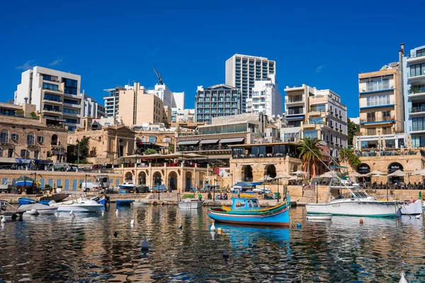 Dalgalanan yansımalar, kireçtaşı atari salonları, modern kuleler, palmiye ağaçları ve gündüz ışığı altında kafe teraslarıyla, Malta, Sliema 'da renkli luzzu tekneleri sallanıyor..