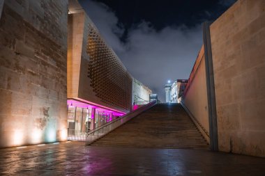 Malta 'daki Valletta Şehir Kapısı' nın gece görüşü Parlamento Binası 'nın delinmiş ön cephesi ve mor zemin katı, geniş merdiveni, ıslak kaldırımı ve devasa taş duvarları olduğunu gösteriyor..