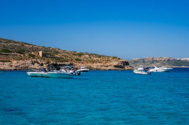 Malta 'da Comino ve Gozo arasında turkuaz mavi gölde boş tekneler yüzüyor. Rocky Sahili ve Gözetleme Kulesi, gün ışığında sığ suları ve geniş manzarayı temizler..