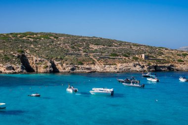 Küçük tekneler ve sürat tekneleri Malta 'nın Comino Adası' ndaki Blue Lagoon 'da dinleniyor. Sığ turkuaz körfez, kayalık kireç taşı kıyısı, çalılık ve parlak ışık altında küçük bir taş yapı..
