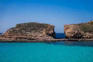 Comino Adası, Malta 'daki Blue Lagoon' daki turkuaz sularda kaya kanalı, kireçtaşı uçurumlar, yaz ışığı ve açık körfezi noktalayan küçük yüzücüler var..