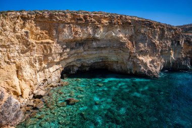 Comino, Malta 'daki bir deniz mağarası etrafındaki kireçtaşı renkli kayalıklar kayalıkların ve resiflerin olduğu sığ turkuaz sulara düşüyor.