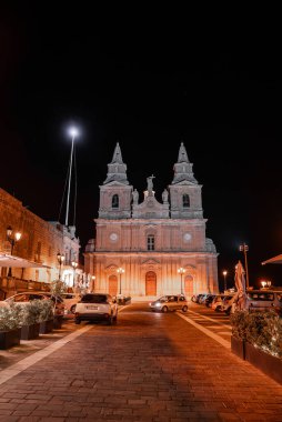 Malta 'daki Mellieha Kilisesi' ne parkedilmiş arabaların kaldırım taşı caddesi hizasında olduğu sıcak ışıklar kondu. Kehribar lambaları parlıyor, ikiz çan kuleleri yükseliyor ve detaylar gece ortaya çıkıyor..