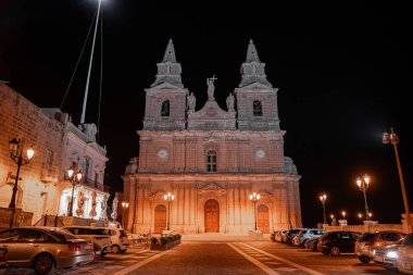 Mellieha Leydisi 'nin mabedi geceleri ana meydanın üzerinde duruyor. Park halindeki arabalar, lamba direkleri ve kemerli kapılar ve borularla döşenmiş kireçtaşı binalar..