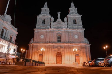 Sıcak kehribar ışıkları, Malta 'nın Mellieha kentindeki plazayı otomobil ve kafe olarak gösteren kumtaşı cephesi, heykel ve kapıları gösteriyor. Düşük açı ve karşıtlık vurgulama masonluğu.