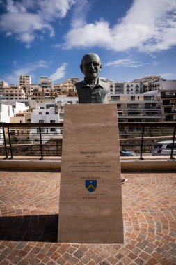 Mellieha gezinti güvertesinde bronz bir büst duruyor. Mavi sarı bir arma, demir korkuluklar, desenli kaldırımlar ve gün ortasında teraslı bloklar..