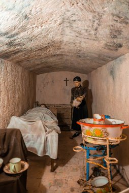 Bir manken hemşire yatağın yanında duruyor. Üzerinde küçük bir Maltese hava saldırısı sığınağı, duvarda 2. Dünya Savaşı sığınağı, mavi bir stantta bebek banyosu, sıcak düşük ışık, dar çerçeve..