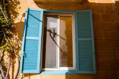 Malta 'nın Mdina kentindeki kireçtaşı bir duvarda turkuaz camlı, beyaz çerçeveli bir pencere açılıyor. Sıcak gün ışığı, sarmaşığın soldan tırmanması gibi kıtır kıtır gölgeler bırakır..