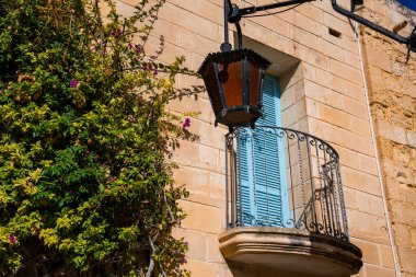 Mdina, Malta 'daki kireçtaşı cephesinde gökyüzü mavisi panjurları ve siyah bir feneri olan küçük dövülmüş demir bir balkon var..