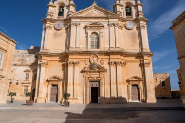 St. Pauls Katedrali, güneşli bir Mdina meydanı, ikiz çan kuleleri, merkezi pediatri ve görülebilir iki saat yüzlü, sıcak kireçtaşı ve dekoratif kapılar görünürde yer alır..