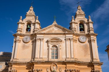 Güneşli Barok cephesi, Mdina, Malta 'da ikiz çan kuleleri, yuvarlak saatler ve yeşil bir haçla yükseliyor. Bal kireç taşı gündüz ışığında parlar, oyulmuş ayrıntıları ortaya çıkarır..