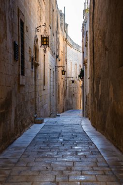 Mdina, Malta 'da dar bir kaldırımlı sokak, kireçtaşı duvarlar arasında rüzgarlar dövmeli demir fenerler ve pencereler arasında, gün ışığıyla aydınlatılan ve eski dokuları gösteren.