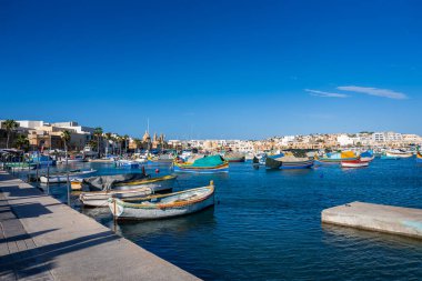Düzinelerce luzzu teknesi Marsaxlokk, Malta 'daki derin mavi sularda sallanıyor, brandalar ve şamandıralar, palmiye çizgili gezinti güverteleri, ve temiz ışık altında kilise kuleleri ve kubbeleri.