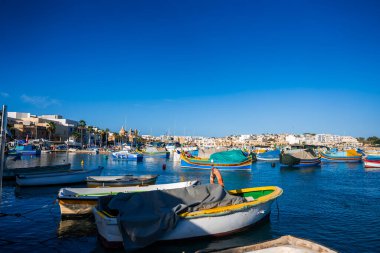 Marsaxlokk, Malta 'da renkli Malta luzzu tekneleri, alçak rıhtımlı binaların yanında yüzer ve berrak öğle ışığında ikiz bir kilise, durgun suya yansıyan canlı boyalar.