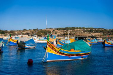 Geleneksel Malta luzzu tekneleri Marsaxlokk, Malta 'da yüzer, canlı renkleri derin mavi suları yansıtır. Öğleden sonra ışığı tepe örtülerini, ağları ve teraslı tepeleri gözler önüne seriyor..