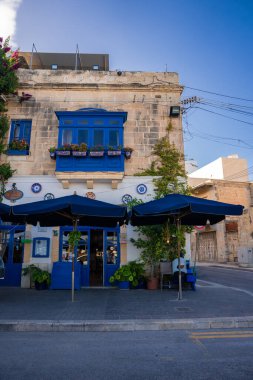 Marsaxlokk, Malta 'da günışığı sokağında kobalt balkonu ve kapıları olan bir kireçtaşı bina, kaldırımda oturan kafe, mavi tenteler, kablolar ve sıcak öğleden sonra ışığı görünüyor..