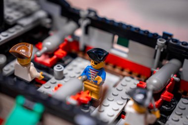 Üç LEGO denizcisi, gri güverte plakalarında duruyor. Kırmızı top arabaları her iki tarafta. Mavi ceketli bir memur önderlik ediyor. Sıcak ışık, sığ odak, oyuncak ölçeği detayı.