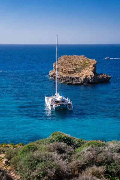 Mavi Lagün, Comino, Malta 'daki kireçtaşı yığınlarının yanında beyaz bir katamaran gün ortasında yüksek bir sahilden kobalt ile su ve yaz ışığı arasında uzanır..