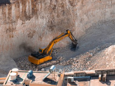 Turuncu bir Hyundai kazıcısı Mellieha, Malta 'daki dik bir kireçtaşı duvarı keserek çatıdaki tankların yakınındaki molozları ve sert gün ışığında tabakları kaldırırken siyah egzoz gazı gönderir..
