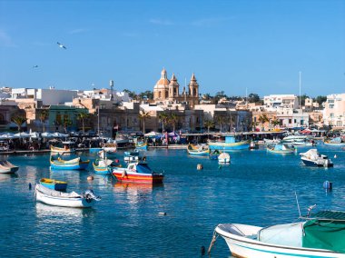Renkli luzzu tekneleri Marsaxlokk, Malta 'da turkuaz bir limanda sallanırken, Pompei Leydimiz Kilisesi' nin kilisesi ve gün ışığında pazar faaliyetleri görülebilir..