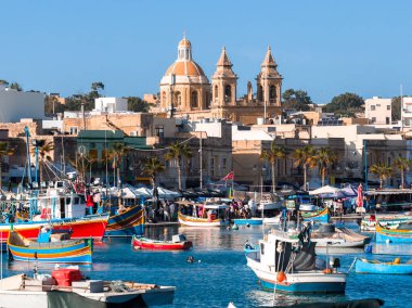 Geleneksel luzzu tekneleri Marsaxlokk, Malta 'da turkuaz suda sallanırken, satıcılar ve ziyaretçiler palmiye tezgahları ve kireçtaşı binaların yakınına, gün ortası güneşinin altında hareket ediyorlar..
