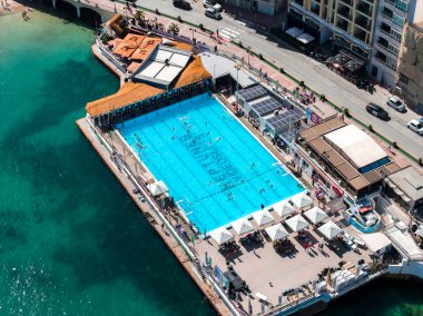 Hava görüntüsü Malta 'daki Sliema yüzme kompleksini gösteriyor. Olimpiyat havuzu, şerit çizgileri, güneş gözlükleri, katlı koltuklar, çadırlar, yüzücüler ve öğle vakti deniz kenarındaki binalar..