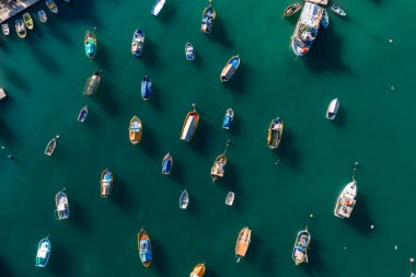 Hava panorama, Marsaxlokk limanını gün ortasında renkli luzzu, küçük botlar ve sakin turkuaz su boyunca uzanan daha büyük çalışma gemileriyle gösteriyor..