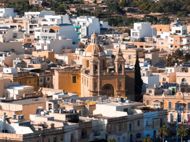 Marsaxlokk, Malta 'nın yüksek manzarası, düz çatılar, güneş panelleri, beyazlatılmış bloklar ve gün ortasında ballı taş evler arasında kubbesi ve ikiz kuleleri olan bir Barok kilisesini gösteriyor..