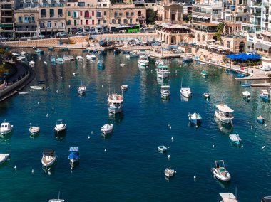 Hava görüntüsü Sliema marinasını turkuaz su, kireçtaşı cepheleri, kemerli atari salonları ve deniz kafeleri üzerinde renkli teknelerle gösteriyor. Yeşil bir otobüs kıyı yolu boyunca ilerliyor..