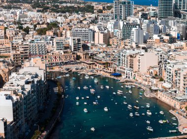 Hava görüntüsü, Sliema St. Julians, Malta 'daki Spinola Körfezi' ni gösteriyor. Hilal bir limanı, küçük beyaz tekneleri, kıvrımlı bir gezinti güvertesi, yükselen daireler ve gün ortası aydınlığı..