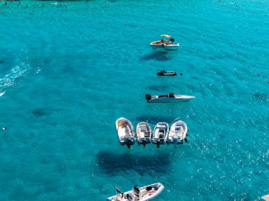 Yüksek açılı hava manzarası, Malta 'nın Comino Adası açıklarındaki Mavi Göl' de küçük tekne ve yüzücüleri gösteriyor. Turkuaz su, aşağıda koyu lekeler, parlak öğle ışığı, sakin yüzey.