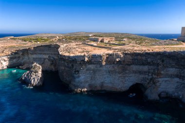 Hava panorama Comino Adası, Malta 'da kireçtaşı kayalıkları gösteriyor. Küçük beyaz bir teknesi olan bir deniz mağarası, Comino Hastanesi kompleksi ve öğle vakti St. Marys Battery..